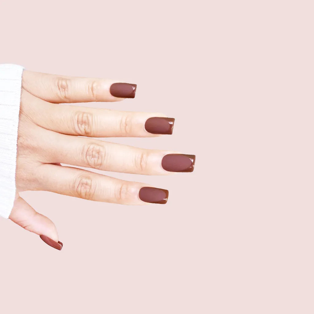 Diagonal hand pose showing de blinkie Cocoa Suede rich chestnut velvet matte press-on nails in medium square shape against pink background with white ribbed knit sleeve